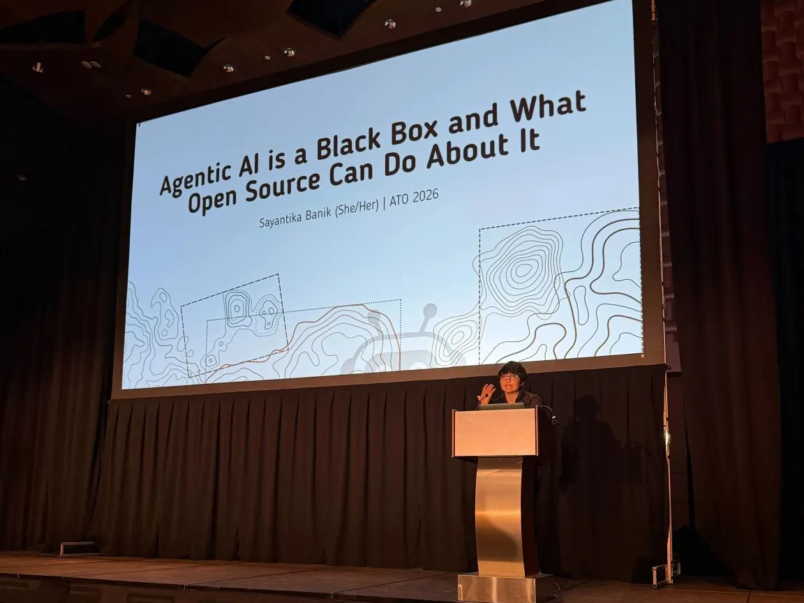 Sayantika Banik speaking at the podium at All Things Open 2026, under the slide "Agentic AI is a Black Box and What Open Source Can Do About It"